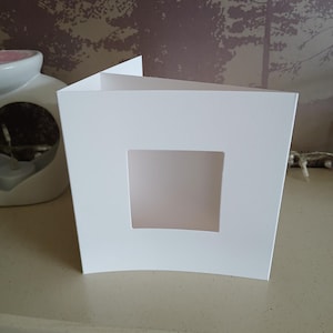 May include: A square white greeting card with a centered square die-cut window. The card is folded vertically and stands upright, featuring a smooth, matte finish on the front panel. The design is simple, clean, and minimalist in its construction.