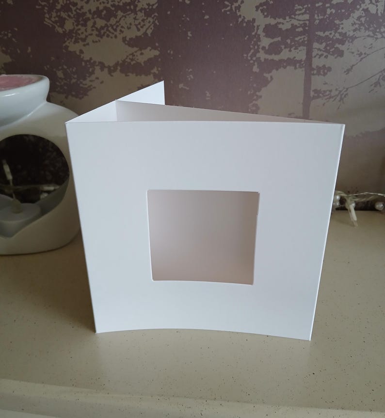 May include: A square white greeting card with a centered square die-cut window. The card is folded vertically and stands upright, featuring a smooth, matte finish on the front panel. The design is simple, clean, and minimalist in its construction.