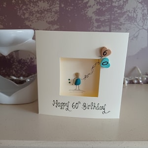 Peut inclure: Une carte faite &agrave; la main avec un design en galets. Un petit personnage tient une cha&icirc;ne de ballons en forme de c&oelig;ur turquoise et marron. La carte indique "Joyeux 60e anniversaire".