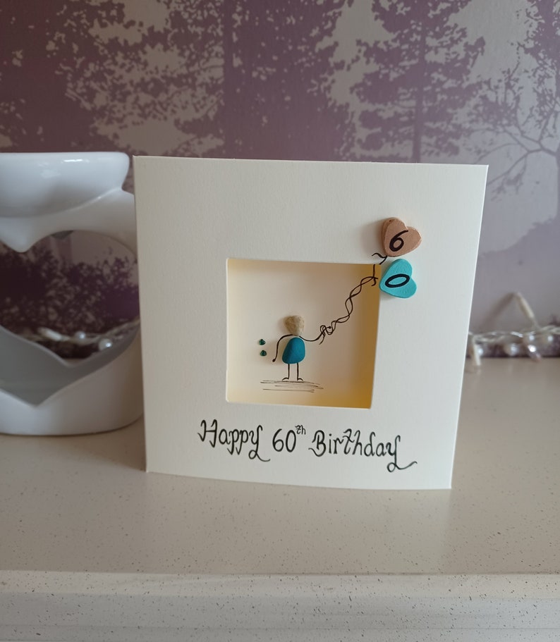 Peut inclure: Une carte faite &agrave; la main avec un design en galets. Un petit personnage tient une cha&icirc;ne de ballons en forme de c&oelig;ur turquoise et marron. La carte indique "Joyeux 60e anniversaire".