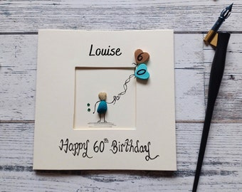 May include: A square greeting card featuring the name Louise and the text Happy 60th Birthday. The card displays a pebble art figure holding two heart-shaped balloons, one brown with the number 6 and one teal with the number 0, next to a pen.