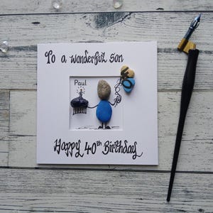 Peut inclure: Carte d'anniversaire faite à la main avec le message "To a wonderful Son" et "Happy 40th Birthday". La carte présente une figurine en galets tenant un cupcake et des ballons. Un stylo de calligraphie est posé sur le côté.