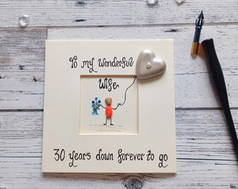 May include: A white wooden frame with a pebble art picture inside. The picture shows a stick figure holding flowers and a heart balloon. The text on the frame reads "To my wonderful wife 30 years down forever to go".