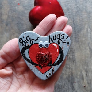 May include: A gray heart-shaped ornament with a red heart inside, googly eyes, and the words "Big hugs!" painted on it.