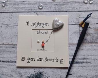 May include: A square greeting card with cream cardstock featuring the text To my Gorgeous Husband and 30 years down forever to go. A pebble art figure in red holds a string attached to a silver heart balloon. A black calligraphy pen sits nearby.