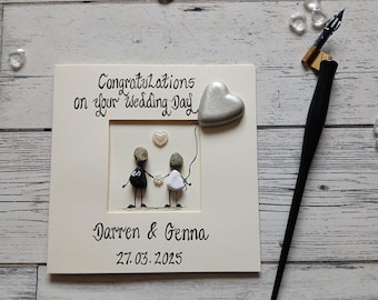 Puede incluir: Una tarjeta de boda color crema con el texto "Congratulations on your Wedding Day" y los nombres "Darren & Genna" junto con la fecha "27.03.2025". La tarjeta presenta arte de guijarros de una pareja tomados de la mano, un globo en forma de corazón y un corazón pequeño.