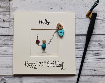May include: A square cream-colored greeting card featuring the name Holly and the text Happy 21st Birthday. The card displays a pebble art figure holding a cupcake with a lit candle and two heart-shaped balloons labeled 2 and 1.