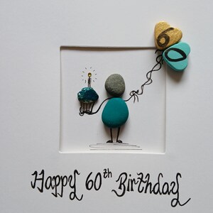 May include: A white greeting card featuring pebble art of a figure holding a cupcake with a lit candle and two heart-shaped balloons with the number 60 written on them. The text Happy 60th Birthday is printed in black ink at the bottom of the card.