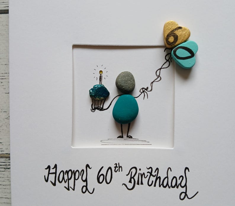 May include: A white greeting card featuring pebble art of a figure holding a cupcake with a lit candle and two heart-shaped balloons with the number 60 written on them. The text Happy 60th Birthday is printed in black ink at the bottom of the card.