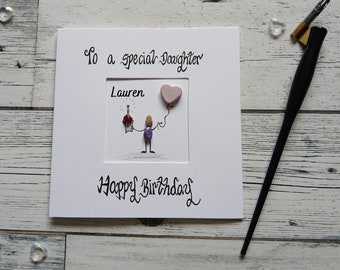 May include: A square white greeting card with the text To a special Daughter and Happy Birthday in black script. The center features a pebble art figure holding a cupcake with a lit candle and a pink heart-shaped balloon, labeled Lauren.
