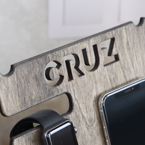 May include: A wooden phone and watch stand with the word "CRUZ" carved into the top. The stand has a slot for a phone and a watch.