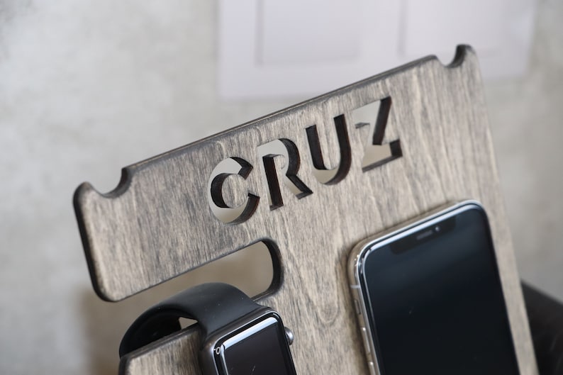 May include: A wooden phone and watch stand with the word "CRUZ" carved into the top. The stand has a slot for a phone and a watch.