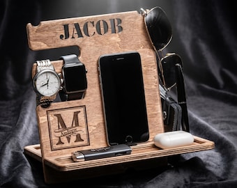 May include: A wooden docking station featuring the name JACOB engraved at the top and a monogram with the name Andrew inside a square frame. The stand holds a smartphone, a wristwatch, a smartwatch, sunglasses, a pen, and a small white case.
