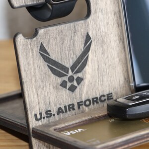 May include: Wooden phone stand with a gray finish. The stand has a slot for a phone, a watch, and a key. It also has a space for a credit card. The stand is engraved with the U.S. Air Force logo and the words "U.S. Air Force".