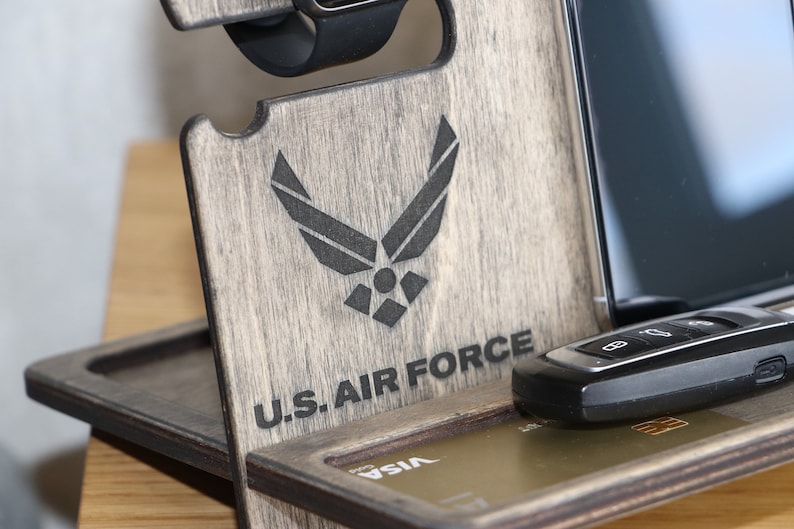 May include: Wooden phone stand with a gray finish. The stand has a slot for a phone, a watch, and a key. It also has a space for a credit card. The stand is engraved with the U.S. Air Force logo and the words "U.S. Air Force".