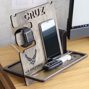 May include: A wooden phone and watch stand with a gray wood finish. The stand has a slot for a phone, a watch, and a small tray for keys and other items. The stand is personalized with the name "CRUZ" and the U.S. Air Force logo.
