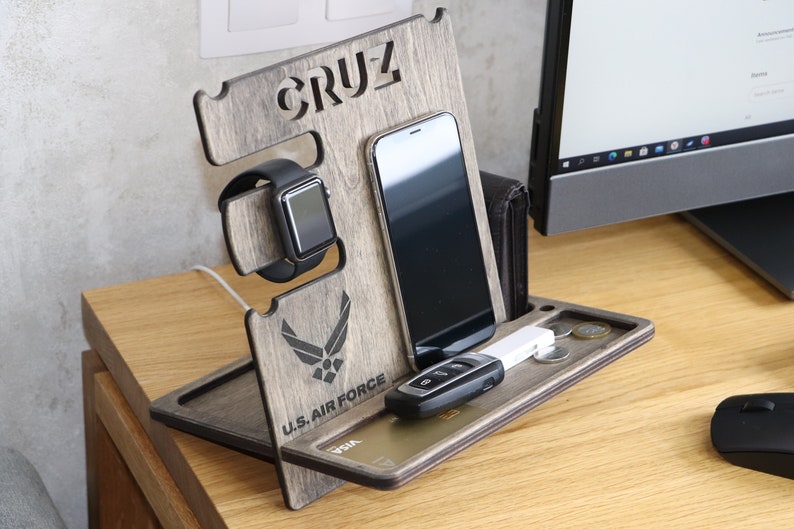 May include: A wooden phone and watch stand with a gray wood finish. The stand has a slot for a phone, a watch, and a small tray for keys and other items. The stand is personalized with the name "CRUZ" and the U.S. Air Force logo.