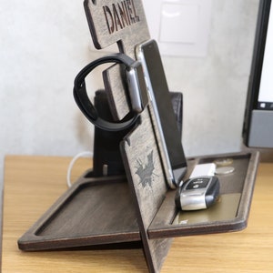 May include: A wooden phone and watch stand with a personalized nameplate that reads "DANIEL". The stand has a slot for a phone, a watch holder, and a tray for keys and other small items. The stand is made of dark wood and has a bat design.