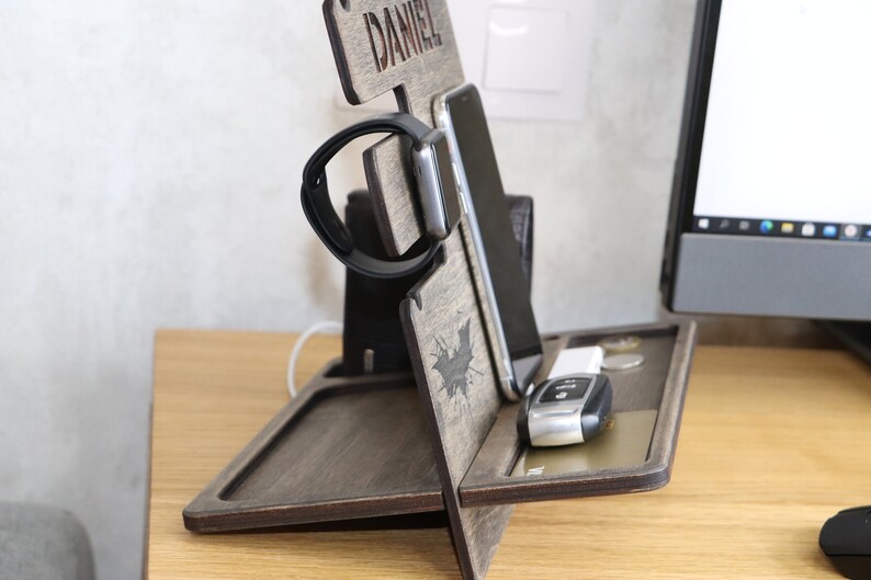 May include: A wooden phone and watch stand with a personalized nameplate that reads "DANIEL". The stand has a slot for a phone, a watch holder, and a tray for keys and other small items. The stand is made of dark wood and has a bat design.