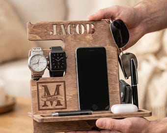 May include: A brown wooden docking station featuring the name JACOB engraved at the top and a monogram with the name Andrew inside a square. The organizer holds a wristwatch, a smartwatch, a smartphone, sunglasses, a pen, and small accessories.