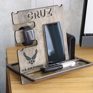 May include: A wooden phone stand with a gray wood finish. The stand has a slot for a phone, a watch, and a space for keys and cards. The stand is personalized with the name "CRUZ" and the logo of the U.S. Air Force.