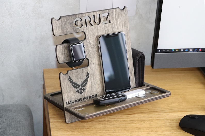 May include: A wooden phone stand with a gray wood finish. The stand has a slot for a phone, a watch, and a space for keys and cards. The stand is personalized with the name "CRUZ" and the logo of the U.S. Air Force.