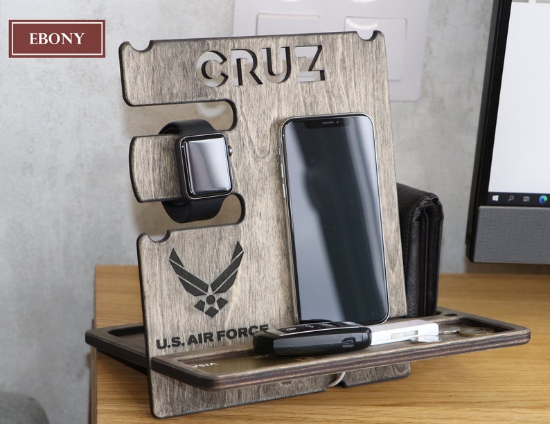 May include: A wooden phone stand with a gray finish. The stand has a slot for a phone, a watch, and a key. It also has a small tray for other items. The stand is personalized with the name "CRUZ" and the U.S. Air Force logo.