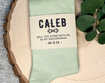 May include: A light green sock with a brown tag that says "Caleb Will You Stand With Me As My Groomsman? - 04.13.24 -" with a bow tie icon.
