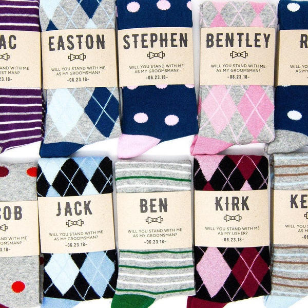 Op de afbeelding: Tien paar sokken met gepersonaliseerde labels. Elke sok heeft een ander patroon, waaronder strepen, ruitjes en stippen. De labels bevatten de namen "Mac", "Easton", "Stephen", "Bentley", "Ross", "Jacob", "Jack", "Ben", "Kirk" en "Kevin". De labels bevatten ook de tekst "Wil je met me staan als..." gevolgd door "getuige", "bruidegom" of "usher" en een datum.