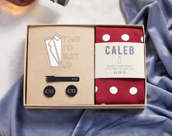 May include: A gift box set for a man with a red and white polka dot sock, a tie clip, cufflinks, and a wooden cutout of a suit with the text "Time to Suit Up!" and the date "03.28.23".