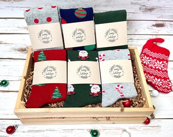 May include: A wooden box filled with six pairs of Christmas themed socks. The socks are wrapped in brown paper with the words "Happy Holidays" printed on each. The box is sitting on a white wooden surface.