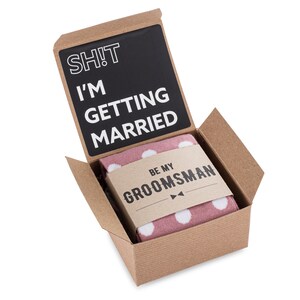 May include: A brown cardboard box with a pink and white polka dot sock inside. The box is open and the sock is visible. The box has a black card inside with the text "SH!T I'M GETTING MARRIED" and a smaller card with the text "BE MY GROOMSMAN".