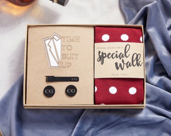 Könnte beinhalten: Eine Geschenkbox aus braunem Karton. Die Box enthält ein Paar rote Socken mit weißen Punkten, eine Krawattenklammer, zwei Manschettenknöpfe und eine Holzaussparung eines Anzugs mit dem Text "Time to Suit Up". Die Socken haben ein Etikett mit der Aufschrift "Special socks for a special walk - love, mari - "