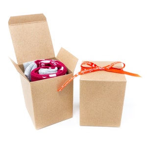 Peut inclure: Deux boîtes cadeaux en carton brun, l'une contenant une paire de chaussettes rouges et grises et l'autre attachée d'un ruban orange avec le texte "Joyeux Noël" imprimé dessus.
