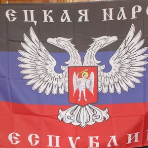 Może przedstawiać: Prostokątna flaga z poziomymi pasami w kolorze czarnym, niebieskim i czerwonym. Flaga przedstawia herb z dwugłowym orłem nad czerwoną tarczą z białą postacią. Tekst cyrylicą jest widoczny u góry i u dołu.