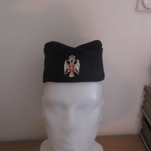 May include: A black military hat, adorned with a silver and red emblem of a double-headed eagle and crown. The hat is displayed on a white mannequin head, highlighting its shape and design. The hat's design suggests a historical or ceremonial purpose.