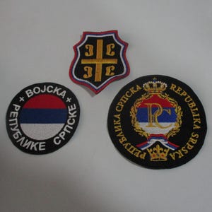 Pode incluir: Três patches bordados com fundos pretos e designs coloridos. O primeiro patch é redondo e apresenta a bandeira sérvia com o texto "Republike Srpske" e "Vojska" em branco. O segundo patch tem a forma de um escudo com uma cruz dourada e as letras "3C3E" em dourado. O terceiro patch é redondo e apresenta uma coroa dourada, uma letra "P" dourada e uma letra "C" dourada com uma coroa de folhas douradas e o texto "Republika Srpska" em dourado.