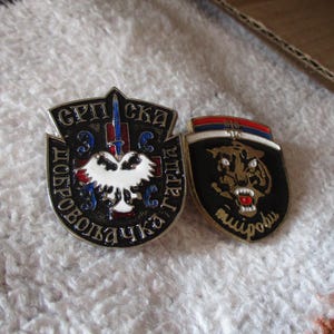 Puede incluir: Dos insignias metálicas. Una es negra con un águila blanca y texto cirílico. La otra es negra con un tigre y texto cirílico, y una franja roja, blanca y azul en la parte superior. Ambas insignias tienen un acabado brillante.