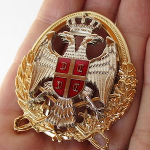 Puede incluir: Un pin de metal dorado con un diseño de águila bicéfala. El águila sostiene un escudo con una cruz roja y cuatro letras blancas. El pin está rodeado por una corona de hojas.