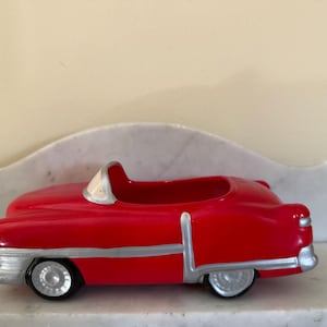 May include: A red ceramic vintage car planter with silver trim and black wheels. The car has an open top and is designed in a classic style. The planter is likely for small plants or decorative use.