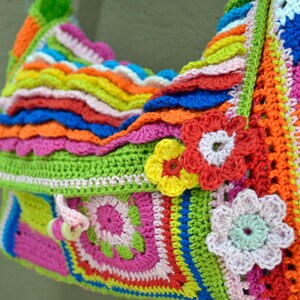 Op de afbeelding: Een kleurrijke gehaakte tas met een verscheidenheid aan felle kleuren, waaronder rood, oranje, geel, groen, blauw en roze. De tas heeft een klep sluiting en een decoratief bloemenontwerp.