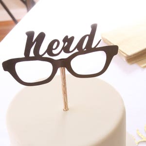 May include: A round, off-white cake with a black "nerd" cake topper featuring glasses. The topper is attached to a wooden stick. The cake sits on a white cake stand, with a white background.