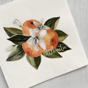 May include: A square paper napkin featuring a watercolor illustration of two orange citrus fruits surrounded by dark green leaves and white blossoms. The text "little cutie" is written in a white cursive script across the lower right side.