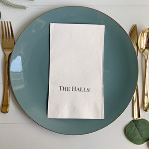 A photo of a beautifully set wedding table with elegant personalized dinner napkins. The napkins feature the bride and groom's first names or last name, printed in a stunning font and surrounded by a delicate decorative border.
