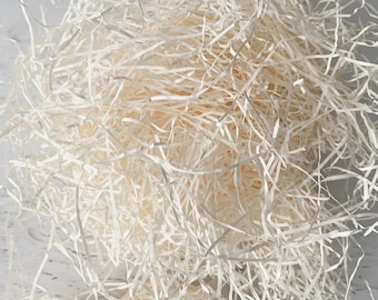 May include: Close-up of a pile of light beige shredded paper, used for packaging and cushioning. The paper is arranged in a dense, tangled mass, with individual strips visible. The background is a light, textured surface.