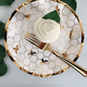May include: White paper plate with a gold honeycomb pattern and gold bee silhouettes. A white meringue cookie sits on the plate with a green leaf on top. A gold fork is on the plate.