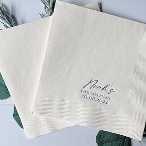 May include: Two white paper napkins with blue text. The top napkin has the text "Noah's Bar Mitzvah 10.08.2022".
