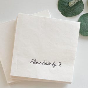 May include: A stack of square, off-white paper napkins featuring the black printed text "Please leave by 9." The napkins are layered slightly offset, showcasing a clean, simple design with a smooth texture and crisp, straight edges.