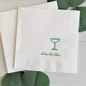 May include: White cocktail napkins with a green margarita glass graphic and the phrase "A tini bit older." The napkins are surrounded by green leaves, suggesting a party or event theme.