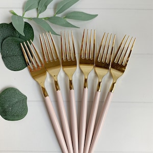 May include: Set of six gold-colored forks with pale pink handles. The tines and upper portion of the forks are gold. The forks are arranged on a white surface, with green leaves in the background.